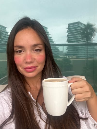 A woman is holding a white mug filled with coffee in front of a window. She has long, dark brown hair, brown eyes, and a fair complexion with red cheeks. She is wearing a white shirt with small floral embroidery. The background shows a modern building reflected in the glass. The building appears to be located in Abu Dhabi, United Arab Emirates.