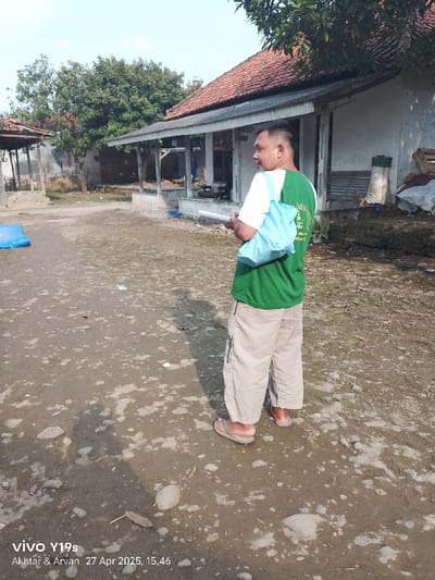 The image shows a man standing in a dirt lot in Anjatan, Indonesia (latitude: -6.419, longitude: 107.979). The man is wearing a green shirt with the words "Satgas" and "Covid-19" on it and beige pants. He is holding a blue bag in his right hand. The man is facing to the left, looking off into the distance. There are several buildings in the background, all of which have red tiled roofs. The ground is muddy and there are several rocks and pebbles scattered about. There is a tree in the background to the left of the man, and another tree in the background to the right. The image was taken on April 27, 2025, at 3:46 PM.