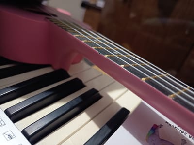 The image shows a close-up of a pink ukulele lying on a keyboard. The ukulele's fretboard is visible, with white strings and golden frets. The keyboard has black and white keys. A piece of paper with a unicorn illustration and Cyrillic text "ЗА ЗАПИСКИ" is underneath the ukulele. The location is Silistra, Bulgaria, but the specific indoor location is unknown.