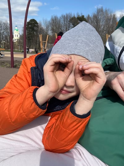 The image captures a child lying down, wearing a gray hat and an orange jacket, with their hands positioned over their eyes. The background reveals a park-like setting, with playground equipment and trees visible. In the distance, the distinctive green and white building of the Church of the Icon of the Mother of God of All Who Sorrow, a landmark in Fryazino, Russia, is identifiable. The overall scene suggests a sunny day in a recreational area.