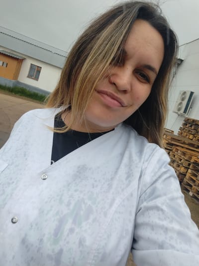 A woman with shoulder-length, layered blonde hair and light brown roots is taking a selfie outdoors. She is wearing a white lab coat with a faint pattern of darker splotches, possibly water stains, over a black t-shirt. A thin silver chain necklace is visible. Her facial expression is relaxed, with her lips slightly parted and her eyes looking towards the camera.

The background suggests an industrial or semi-industrial area. To the left is a low-rise building with a light-colored facade, a yellow-orange trim, and a gray metal roof. A single window with brown framing is visible on the building. Green grass and some dirt are present in front of the building.

To the right of the woman, a stack of wooden pallets is visible, suggesting a storage or loading area. Above and behind the pallets, a white outdoor air conditioning unit with two circular vents is mounted on a white wall. The sky is overcast and light gray, indicating a cloudy day. The overall scene is neutral and lacks any specific identifiers of the city of Foki, Russia, beyond what is implied.