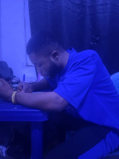 A man wearing a blue shirt and black pants sits on a blue chair and bends forward. He holds a blue pen in his hand, seemingly writing or drawing. He wears a yellow wristband with writing on it. The scene is lit with a strong blue light. A wall is behind him, and what looks like a volleyball is above him.