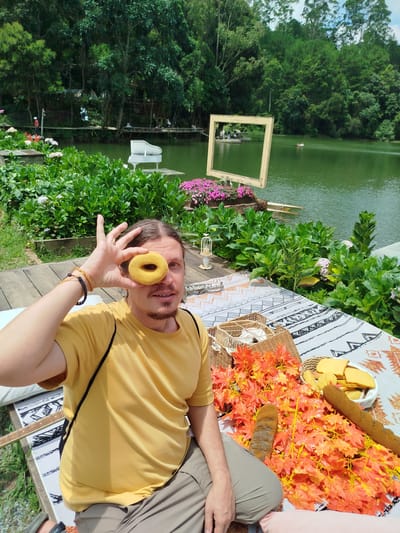 A man sits on a wooden deck next to a lake, holding a yellow donut up to his right eye. He is wearing a yellow t-shirt and khaki shorts. A picnic blanket with a patterned design is spread out on the deck in front of him, adorned with faux autumn leaves, a basket, two baguettes, and cookies. The background features a serene lake surrounded by lush green trees and foliage. In the distance, across the lake, a white grand piano sits on a small platform, and people can be seen on a wooden walkway and in small boats. The overall scene suggests a leisurely picnic setting in a natural environment, possibly at Cam Ly waterfall area in Da Lat, Vietnam, known for its picturesque landscapes and attractions.
