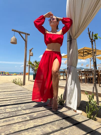 A young woman stands on a wooden boardwalk on a beach, wearing a red off-the-shoulder crop top and a flowing red skirt with a high slit. She has her hands behind her head, looking up at the sky. The boardwalk leads to the beach, where there are inflatable water toys in the ocean. In the background, there is a beachside restaurant with tables and chairs under yellow umbrellas. The scene is set in Quy Nhon Ward, Vietnam, under a clear blue sky.