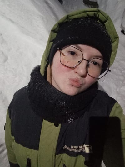 A young female individual is depicted in a close-up, chest-level portrait taken outdoors in a snowy environment in Старый Шагирт, Russia. She is facing the camera directly with pursed lips. Her cheeks exhibit a flushed appearance. She is wearing black-framed, geometric eyeglasses. Her head is covered by a black knit beanie, over which a dark green jacket hood is pulled up. A black, textured neck warmer is worn, showing small white particles consistent with snow. Her jacket is dark green and black, with a central black panel displaying faint, mirrored text and a triangular logo. The background consists entirely of soft, indistinct white snow under dim ambient lighting.