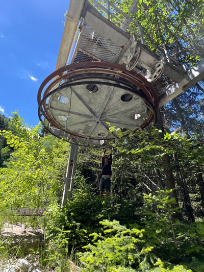 A low-angle shot captures a person hanging from a dismantled ski lift structure amidst a dense forest on a sunny day. The ski lift's large pulley mechanism, rusted and weathered, dominates the upper portion of the frame, with a person, partially visible and silhouetted against the bright sky, hanging from it with their arms extended upwards. The surrounding environment is lush with green foliage, consisting of trees and undergrowth, typical of a mountainous or forested area. The sky is clear blue with scattered white clouds. The scene appears to be located in a rural or natural setting, likely in the vicinity of the city of Samokov, Bulgaria, given the presence of a ski lift, which are common in mountainous regions like the Rila Mountains near Samokov. The overall impression is one of adventurous exploration or playful interaction with abandoned infrastructure in a natural landscape.