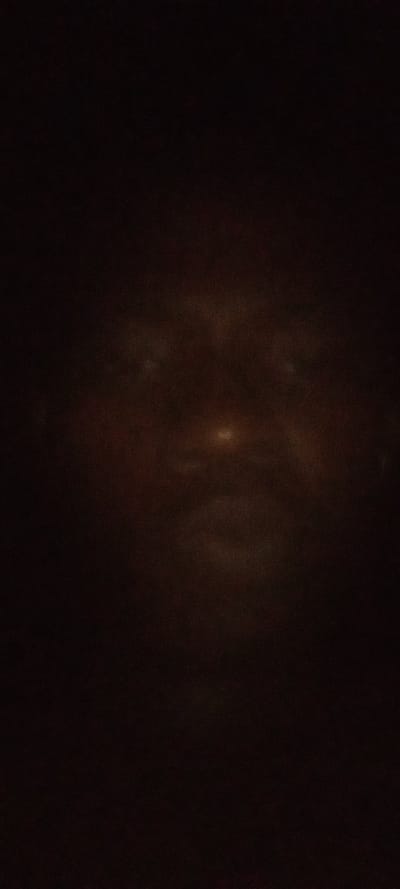 A dark image reveals a faint, ghostly face. Two eyes are subtly visible at the top, along with a nose that is slightly illuminated. Hints of a mouth can also be seen lower down. The face is symmetrical and shadowy against a very dark background.