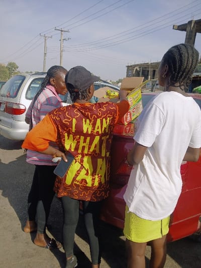 An outdoor daytime scene in Kwekwe, Zimbabwe, depicts three individuals gathered around two parked vehicles. On the left, partially visible, is the rear of a silver minivan. Standing barefoot beside this vehicle is a person with braided hair, wearing a pink-and-white striped shirt and dark leggings, holding a dark flip-flop. In the center, facing away from the viewer, a person in a dark baseball cap, an orange-sleeved, reddish-brown shirt with yellow text partially reading "I WANT WHAT I WANT," and dark leggings, holds a smartphone in their left hand. This individual is actively reaching into an open cardboard box containing yellow-packaged goods, likely instant noodles, which is placed on the trunk of a red sedan. To the right, another person, also facing away, wears a white t-shirt and a bright yellow mini-skirt, standing adjacent to the red sedan. Overhead, multiple utility poles with power lines are visible against a bright, clear sky. In the distant background on the right, a partially constructed building with exposed concrete framework can be seen. The ground is paved asphalt.