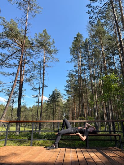The image, oriented vertically, depicts a young woman standing on a wooden deck with a metal railing. She is wearing a black t-shirt with a colorful graphic and black pants, with black sneakers on her feet. Her hair is pulled back, and she is looking to her left with her left hand resting on her hip and her right hand near her forehead. The deck appears to be part of an elevated structure, possibly a balcony or platform, overlooking a dense forest of tall pine trees. The trees are characteristic of the Komarovo, Russia location, with their slender trunks and green needles.
The background is dominated by a clear blue sky, with a few wisps of white clouds visible. The lighting suggests it is daytime, likely mid-morning or afternoon, with the sun casting distinct shadows on the wooden deck. The overall scene conveys a sense of calm and relaxation, with the woman appearing contemplative or enjoying the natural surroundings. The forest setting is lush and green, indicating a warm season. The wooden texture of the deck is prominent, showing the grain of the wood. The metal railing provides a modern contrast to the natural environment. No visible text is present in the image.