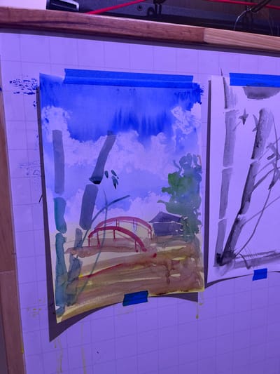 The image depicts two watercolor paintings mounted side-by-side on a white, gridded board in an indoor setting within Bogota, Capital District, Colombia.

The painting on the left is a landscape oriented vertically. Its upper portion features a deep blue sky with white, cloud-like elements, from which blue paint has visibly dripped downwards. The mid-ground includes multiple grey-green vertical forms resembling bamboo stalks, some with small green foliage. A red arched bridge is centrally positioned in the mid-ground, leading to a dark-roofed structure partially obscured by green tree-like shapes on the right. The foreground is composed of various earthy tones, including yellow, brown, and red washes. Green and yellow-brown paint drips extend from the bottom edge of this painting. It is secured to the board with three strips of blue painter's tape: one on each top corner and one at the bottom center.

The painting on the right is a vertically oriented monochromatic study, executed in grey and black tones on white paper, depicting bamboo stalks with angular branches and small leaf-like forms. This artwork appears less detailed than the left painting. It is secured to the board with blue painter's tape strips at the top and bottom.

The white gridded board exhibits minor blue paint splatters to the left of the first painting and yellow-brown paint drips from the bottom of the first painting onto the grid. Above the board, a wooden frame component is visible, along with a red cable and a black mechanical object with a screw. The entire scene is illuminated by a strong purple-blue light, which casts shadows beneath the paintings. No individuals are present in the scene.