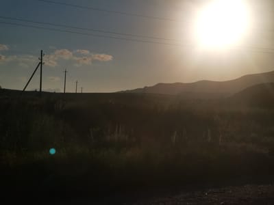 The image depicts a silhouetted landscape under a bright, hazy sun. The foreground is dominated by dark, out-of-focus vegetation, with a small, glowing cyan circle visible in the lower left quadrant. Several utility poles with wires stretch across the frame from left to right, silhouetted against a pale blue sky with a few wispy clouds. In the background, rolling hills are visible as dark shapes against the bright horizon. The scene appears to be outdoors, possibly a rural or semi-rural area, with no discernible human activity or specific landmarks identifying it as being in the city of Kubina, Russia. The overall impression is one of natural scenery at sunset or sunrise, with atmospheric haze obscuring finer details.