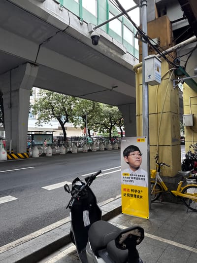 An outdoor, street-level, medium-shot photograph captures a section of an overpass in Guangzhou's Yuexiu District, China. The concrete structure of the overpass dominates the upper portion of the image, showing signs of weathering and a prominent crack. Below the overpass, a street runs parallel to the sidewalk. Several white, concrete bollards line the edge of the sidewalk, separating it from the road. A black electric scooter is parked on the sidewalk in the foreground, with a white surgical mask hanging from its handlebars.
To the right of the scooter, a tall, yellow advertising poster is propped against a concrete pillar. The poster features a portrait of a young boy wearing glasses, with Chinese text overlaying the image. The text appears to be an advertisement for Kodak Lenses, promoting myopia control. To the right of the poster, a yellow bicycle, also a shared bike, is visible. Beyond the sidewalk and to the left, a row of parked bicycles or scooters is partially obscured by the overpass pillars. People are faintly visible in the background, near what appears to be a bus stop or waiting area. The overall scene depicts an urban environment with typical street furniture and infrastructure.