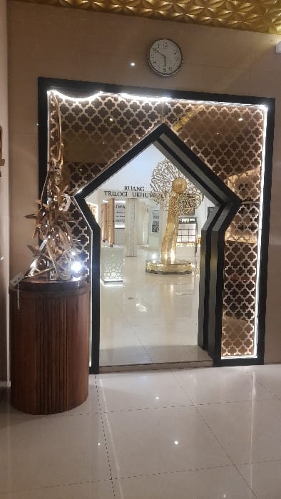 The image depicts an ornate interior entrance within a building located in Glantengan, Indonesia. The central feature is a large archway with a black frame and decorative gold-colored panels featuring a repetitive quatrefoil pattern. White LED strip lighting illuminates the inner perimeter of the archway. Above the archway, on a plain light beige wall, a circular analog wall clock displays approximately 11:45. The ceiling is clad with gold-colored, textured panels forming a geometric pattern. To the left of the archway, a fluted, cylindrical wooden pedestal supports a complex, abstract sculpture composed of multiple interlocked, metallic-hued components, also illuminated by integrated lights. The floor throughout the visible area consists of reflective, light-colored square tiles. Through the archway, a brightly lit interior space is visible. A sign on a white wall in the background reads "RUANG TRILOGI UKHUWAH". Further into the room, a large, golden, abstract sculpture, possibly resembling a stylized tree or figure, stands on an illuminated, ornate circular base. No individuals are present in the scene. The environment appears meticulously maintained and designed, suggesting a public, possibly cultural or commercial, setting.