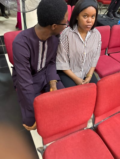 In an indoor setting in Lugbe, Nigeria, a woman with a black headband, wearing a striped brown and white blouse, sits on a red chair. A man wearing glasses and a purple long-sleeved traditional shirt sits next to her. Both are wearing gold bracelets. Several other empty red chairs are visible in the background. A silver fan stands nearby. The floor is black and white tile.