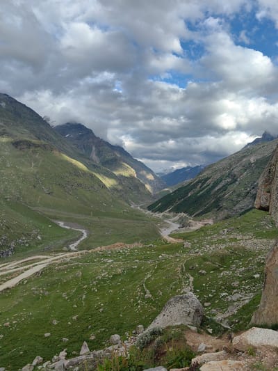 The image depicts a panoramic view of a valley in Gramphu, India. The terrain is mountainous and covered in lush green grass, with scattered rocks and boulders. A winding river flows through the valley, and a dirt road or path is visible alongside it. The sky is filled with dramatic, cloudy formations, with patches of blue sky peeking through. Sunlight intermittently breaks through the clouds, casting illuminated swathes across the slopes. The overall impression is one of a remote, rugged, and beautiful natural landscape. There are no people visible in the image.