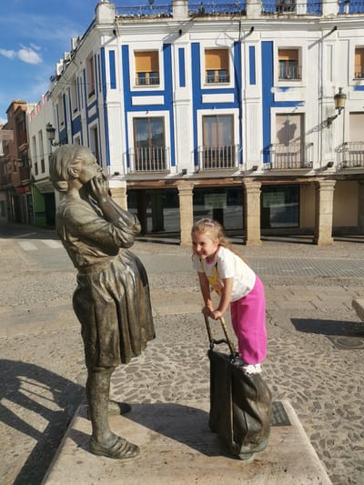 A full shot shows a bronze statue of a woman standing next to a young girl on a stone cobbled street. The statue is of a woman with her hands up to her face, and she is wearing a dress. Next to her, a girl stands atop a bronze sculpture of a suitcase. She is wearing a white shirt, pink pants, and white shoes. She has light brown hair. She smiles as she leans on the handles of the suitcase. Behind them is a building with white and blue paint. The image appears to be taken in the city of Valdepeñas, Spain. The statue is the statue of Agustina of Aragon, on the Plaza de España in Valdepeñas. Under the arched entrance to the building behind the statue are windows with signs posted that say "SE ALQUILA" and phone numbers.