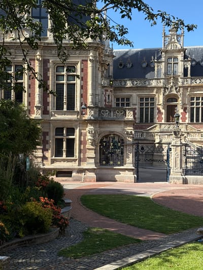 This image captures an ornate historic building, likely a grand château or palace, set against a clear blue sky on a bright, sunny day. The scene suggests a well-maintained historical site or museum, with landscaped grounds in the foreground.
The central subject is the multi-story building, distinguished by its intricate architecture. The facade features a striking combination of light-colored stone and reddish-brown brick arranged in decorative patterns. Numerous windows of various sizes and shapes, some with multiple panes, are visible across the building's face. The structure is adorned with elaborate stone carvings, balustrades, and decorative elements, giving it a rich, gothic or renaissance revival appearance. A prominent dark-tiled roof with several dormer windows and smaller circular openings adds to its grandeur. Below the main windows, decorative metal grates, one featuring star-like motifs, are visible.
In the foreground, lush green foliage from trees and shrubs partially frames the building on the left and bottom right. These plants cast long shadows across the facade and the ground, indicating the strong sunlight. To the right, landscaped grounds feature a winding pathway made of reddish brick pavers, contrasting with a cobblestone path and sections of neatly mown grass. Flowering plants and hedges line the paths and the base of the building.
The bright lighting suggests it is daytime, with clear weather conditions. No people are visible, and no specific activity or event is taking place, presenting a serene and static view of the architectural masterpiece and its surroundings. A small, partially obscured green sign with white, illegible text is visible among the foliage on the right side.
