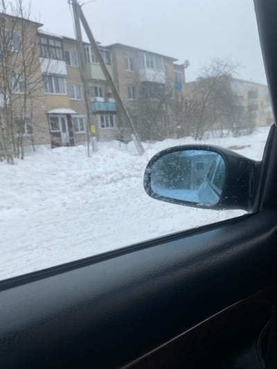 The scene, viewed from inside a vehicle in Карабоўскі сельскі Савет, Belarus, depicts a heavily snow-covered residential area under an overcast sky. The lower right foreground shows the black interior door panel and a black, blue-tinted side mirror, which is spattered with frozen precipitation. The ground is entirely covered by deep, uneven snow, indicating a path or unplowed area. A prominent, leaning wooden utility pole with overhead wires stands in the midground, partially obscuring a multi-story apartment building constructed from light-colored brick or concrete. This building features several balconies; some are enclosed with glass, while others are open, including one painted light blue and another light green. A ground-level entrance includes a small, enclosed porch with a reddish door. A second similar apartment building is visible further in the background. Sparse, bare deciduous trees and bushes are present throughout the snowy landscape. No individuals are visible.