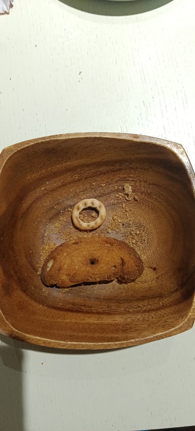 A close-up, overhead shot shows a wooden bowl filled with crumbs and two pieces of bread. A thin, circular cracker with a hole in the center sits above a flat, oval-shaped piece of bread with a hole in its center. Both bread pieces are light brown and appear to be dry and toasted. The bowl is oval and made of dark wood with visible grain. The surface the bowl rests on is a pale green, textured surface, likely a table. The scene is indoors, possibly in a kitchen or dining area, within the Падлабенскі сельскі Савет region of Belarus.