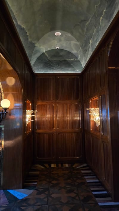 This is a vertical, medium shot captured inside what appears to be a hotel hallway or interior passage. The hallway is narrow, with high, arched ceilings that are painted with a marbled, greenish-blue design, resembling clouds or waves. Two circular, white ceiling lights are visible in the ceiling, along with a smaller, dark security camera.
The walls are paneled with dark wood, featuring vertical grooves within rectangular sections. On either side of the hallway, at mid-height, are illuminated wall sconces with crystalline, wing-like designs that cast a warm glow. To the left, a larger, spherical light fixture with a frosted glass shade is also visible, mounted on a dark, ornate fixture. The floor is a complex mosaic of geometric patterns in various shades of wood and stone, creating a visually striking effect. At the end of the hallway, there is a large, dark wooden door with similar vertical paneling. The overall ambiance is dimly lit, luxurious, and somewhat theatrical.
The location is identified as the Hotel del Coronado, in Coronado, California, which is part of the San Diego metropolitan area, United States of America.