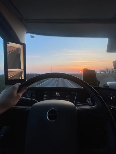 The image presents a first-person perspective from the driver's seat of a Volvo heavy goods vehicle, traveling on a paved two-lane road. The scene is set during either sunrise or sunset, with the sun, appearing as a distinct orange orb, low on the horizon ahead. The sky transitions from a light blue overhead to shades of pink and orange near the horizon.
In the foreground, a driver's right hand is positioned at the top of the dark, multi-function steering wheel, which bears the "VOLVO" logo. The digital instrument cluster behind the steering wheel displays "82," indicating a speed of 82 km/h, along with other vehicle information. To the left of the windshield, a vertical digital display, functioning as a digital mirror system, shows two separate camera views of the road. Another electronic device, likely a navigation unit or smartphone, is mounted on the dashboard to the right of the steering wheel.
The road ahead is straight and recedes into the distance, flanked by a flat landscape. Sparse trees are visible along the right side of the road, and overhead, multiple power lines stretch across the sky, supported by utility poles in the mid-distance. The environment appears rural, consistent with areas around Slaný, Czechia.