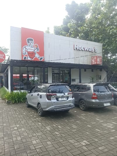 The image shows the exterior of a "Hotway's" restaurant in Purwokerto, Indonesia. The restaurant is located at coordinates -7.418, 109.251, and is a single-story building with a white facade. There is a large sign above the entrance with the restaurant's name and a cartoon image of a man holding a drumstick. The exterior walls are white and there are large windows visible. Two silver cars are parked in front of the restaurant. There is a sign on the right side of the building advertising "Hot Hot" chicken. There are green plants in front of the restaurant and a paved area for parking.