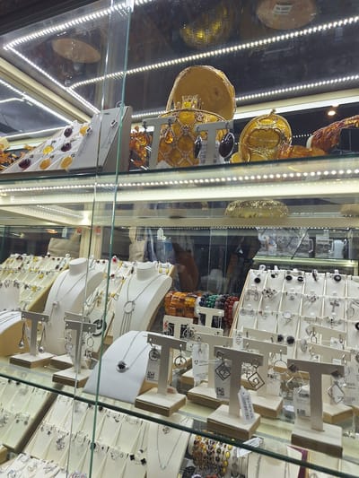 The image shows a display of jewelry inside a brightly lit glass case, likely a retail store. The jewelry includes necklaces, earrings, and rings, many of which appear to be made of silver and feature gemstones or amber. The display is organized on white stands and layered shelves. In the background, other items are visible, including what appears to be amber jewelry and decorative objects with a golden sheen. The lighting is artificial and comes from LED strips lining the case. Reflections in the glass show the store environment, including more displays and possibly other customers. There is no clear indication of time of day or weather, but the indoor setting suggests it could be any time. There are no people clearly visible in the foreground. Some labels are visible on the jewelry and stands, but the text is too small to be decipherable. The overall impression is a well-stocked jewelry shop. Given the context of Peterhof, Russia, this could be a shop selling souvenirs or local crafts.