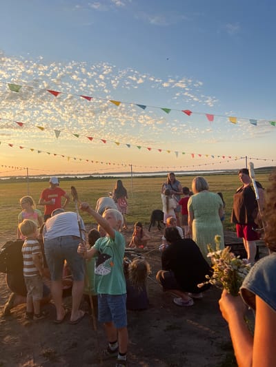 The image depicts an outdoor gathering with people of various ages in a grassy field near the Am Salzhaff, Germany, indicated by the distant body of water and landscape. A string of colorful triangular flags is strung across the sky, set against a backdrop of a soft orange sunset and scattered white clouds. In the foreground, a group of individuals, including children and adults, are gathered around what appears to be a fire pit or a gathering spot. Some individuals are holding sticks, possibly for roasting food. One child in the foreground is wearing a green t-shirt with a fish graphic and blue shorts, holding a stick. Another boy in a striped shirt and shorts stands nearby. Adults are seen standing and sitting, with one woman in a green dress and another in a dark top and red shorts. The ground is composed of dirt and grass. The overall atmosphere suggests a casual outdoor event, possibly a picnic or a campfire gathering.