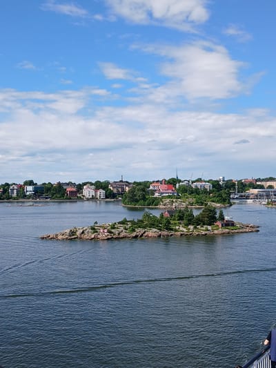 The image depicts a scenic view of Helsinki, Finland, likely taken from a ferry or boat. In the foreground, the dark blue water of the sea is textured with gentle waves and the wake of a vessel. A small, rocky island with sparse green vegetation sits in the middle ground, featuring two small red buildings.

Beyond the island, the coastline of Helsinki is visible, characterized by a dense arrangement of trees and a variety of buildings. These buildings range in color and architectural style, with some appearing to be residential homes and others more institutional or commercial structures. Prominent red-roofed buildings are interspersed among white and muted-colored facades. Several sailboats are docked along the shore, indicating a harbor or marina. Cranes and towers are also visible on the mainland, suggesting ongoing construction or industrial activity.

The sky above is a vibrant blue, dotted with white, fluffy cumulus clouds. The lighting suggests it is daytime, with the sun casting a clear light on the scene. The overall impression is one of a pleasant day in a coastal city, with a mix of natural beauty and urban development.