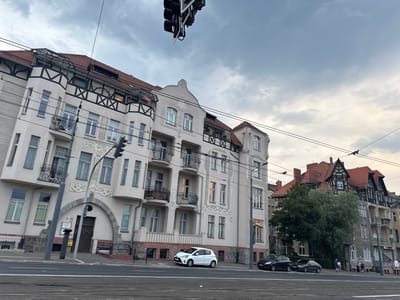 The image shows a view of a street in Poznan, Poland, with several buildings and cars. The architecture suggests an older European city. In the foreground, there is a street with tram tracks, and a white car is parked on the side. Behind the car, two black cars are also parked. A few people are walking on the sidewalk. The buildings have ornate facades with multiple floors, balconies, and red-tiled roofs. A traffic light is visible in the upper part of the image. The sky is overcast. The specific location appears to be near the intersection of Ratajczaka Street and Gwarna Street in Poznan, based on the architectural style and the presence of tram tracks.