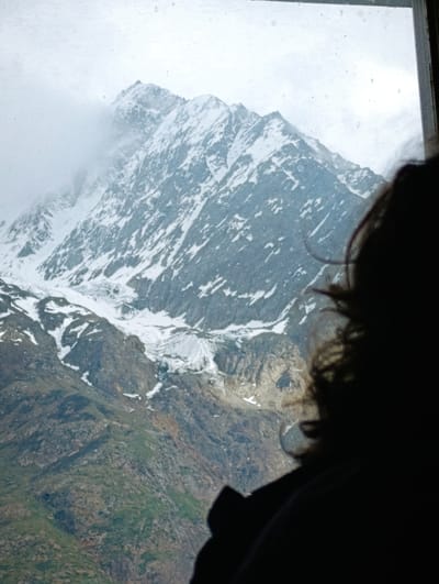 The image shows a view of snow-capped mountains through a window, with the silhouette of a person's head and shoulders in the foreground obscuring part of the view. The mountains are rugged and rocky, with patches of snow and ice. Clouds are visible around the peaks. The lower slopes of the mountains appear to be covered in green vegetation and rocky terrain. The overall lighting is dim, suggesting an overcast or early morning/late evening scene. The location is identified as Gemur, India, a place known for its mountainous terrain.