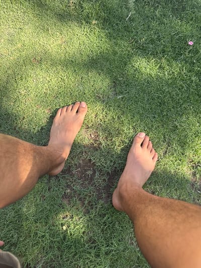 The image shows a pair of bare feet on a grassy lawn. The feet are positioned close to each other, with the left foot slightly in front of the right. The grass is green and appears to be well-maintained, with some patches of sunlight filtering through. The overall scene suggests a casual, outdoor setting, possibly a park or backyard. The lighting indicates it is daytime, likely late morning or early afternoon, with bright sunlight creating shadows on the grass. There are no visible signs of text or specific landmarks to pinpoint the exact location within El Paso, United States of America, beyond the general context provided. The image evokes a sense of relaxation and connection with nature.