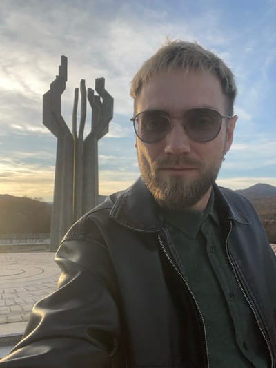 An image taken in Barutana, Podgorica, Montenegro, depicts a male subject in a close-up selfie format, positioned in the right and central foreground. The male, appearing approximately 30-40 years old, has short blond hair and a neatly trimmed blond beard. He wears aviator-style sunglasses with dark lenses, a dark green collared shirt, and a black leather jacket. A silver stud earring is visible in his left earlobe. His gaze is directed forward towards the camera, maintaining a neutral expression.

Behind the subject, centrally located in the mid-ground, stands a large, multi-component concrete monument. This structure, identifiable as the Monument to the Partisan Fighter, consists of several tall, abstract, angular pillars that extend upwards, suggesting a stylized, open hand or flame. The monument is situated on a flat, paved circular plaza featuring geometric patterns.

The background reveals a natural landscape with distant rolling hills, sparsely covered with vegetation. The sky is partially cloudy, displaying shades of light blue, white, and orange-pink near the horizon, indicative of late afternoon or early evening. The sun, positioned low and to the left, casts a warm, golden light across the scene, highlighting the monument and parts of the plaza.