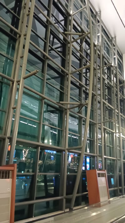 This is an interior shot of a large, modern building with a prominent glass and metal facade. The structure appears to be an airport terminal or a large public space. The architecture features a complex system of silver-toned metal beams and supports that create a geometric lattice against large panes of green-tinted glass. Through the glass, reflections and distant lights suggest an external environment, possibly a cityscape or an airport tarmac at night.
Two orange, rectangular structures with a grated surface are visible in the foreground and mid-ground, suggesting seating or ventilation units. The overall lighting is dim, with artificial light sources creating highlights and reflections. The image's perspective is slightly angled upwards, emphasizing the scale and complexity of the architectural design.
The image is specifically identified as being taken in Tangerang, Indonesia, which is a city known for its proximity to Soekarno-Hatta International Airport, a major transportation hub. The architectural style is consistent with modern airport infrastructure found in large international cities.