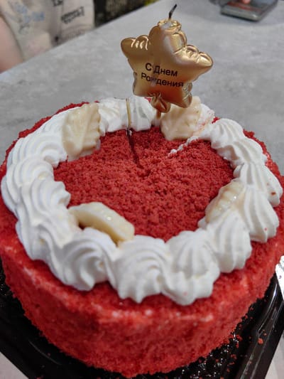 The image shows a close-up of a red velvet cake decorated with white frosting and some pale, leaf-shaped garnishes. A gold star-shaped candle with Cyrillic text that reads "С Днем Рождения" (Happy Birthday) is stuck into the top of the cake. The cake is placed on a black plastic cake stand. The background is blurred, showing a grey, textured surface, possibly a table, and a hint of someone's arm and clothing. The scene appears to be indoors. There is no discernible information within the image that suggests a specific location in the city of Fanipal, Belarus.