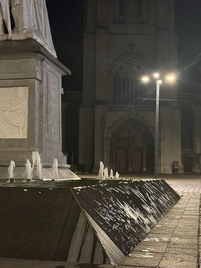 The image depicts a nighttime scene in front of Saint Bavo's Cathedral in Ghent, Belgium. A large stone monument with bas-relief sculptures stands in the foreground on the left. In front of the monument, a fountain with several spouts of water is visible. The water flows down a dark, angled surface, reflecting the light from street lamps. The cathedral facade, characterized by Gothic architecture with a large arched entrance and stained-glass windows, is visible in the background. A street lamp with three lights stands near the center of the image, illuminating the area. The ground is paved with stone. No people are present in the frame.