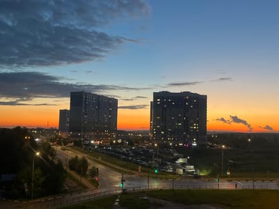 This media file depicts an urban scene at dusk in Domodedovo, Russia. The foreground shows a road with cars and streetlights, indicating it's a busy area. In the midground, several tall, modern apartment buildings are visible, their windows illuminated, suggesting that people are home. A large parking lot filled with cars is situated between the buildings, hinting at a residential complex. The background features a landscape of trees and distant structures, with a dramatic sunset painting the sky in vibrant hues of orange, yellow, and blue.
The time of day is clearly dusk, with the fading sunlight casting long shadows and the sky transitioning from daylight to night. The presence of streetlights and illuminated building windows confirms this. The weather appears clear, with some scattered clouds visible in the upper sky, catching the sunset's colors. The scene evokes a sense of tranquility and the end of a day, with the urban environment set against a beautiful natural phenomenon. There is no visible text in the image.