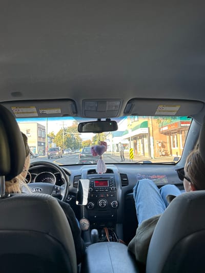 The image depicts the interior of a Kia vehicle, viewed from the rear passenger seat, looking forward through the windshield onto Hillside Avenue in Victoria, British Columbia, Canada. The perspective is directed west from the vicinity of Quadra Street.
In the driver's seat, a person with light-colored hair and earrings is visible from behind, hands on the steering wheel. To their right, a front passenger is seated with their back to the camera, wearing blue jeans and an olive-colored top, with their feet elevated and resting on the dashboard. Dark sunglasses are present on the passenger's head or face.
The vehicle's dashboard and central console are visible, featuring a digital display with red text, climate controls, and a gear selector. A phone holder is affixed to the dashboard to the left of the console, and a circular "SEX WAX" air freshener hangs below it. A decorative item consisting of two purple heart shapes and a white tassel hangs from the rearview mirror, which also has a small camera attachment. Warning labels are visible on the sun visors.
Through the windshield, Hillside Avenue stretches into the distance. A dark-colored vehicle is directly ahead in the lane of travel. Further along the road, a reddish-brown pickup truck and other vehicles are present. On the right side of the street (north side of Hillside Avenue), commercial buildings are visible. One building features a green awning, and an adjacent building displays a red sign reading "BARBER SHOP" with "PASTRIES" visible above it. A pedestrian, dressed in light-colored clothing, stands on the sidewalk near the barber shop, facing away from the camera. Utility poles and overhead wires are also visible. On the left side of the street (south side of Hillside Avenue), various buildings are present, with trees in the background. The scene is illuminated by daylight under a clear to partly cloudy sky.