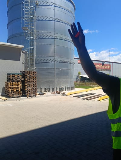 The image shows an industrial area with a large, metallic silo in the background, under a bright blue sky with scattered clouds. In the foreground, a person wearing a high-visibility yellow vest is partially visible, with their arm raised and holding a small, orange object. Several stacks of wooden pallets are arranged on the paved ground to the left of the silo. In the middle ground, there are some metal beams and yellow barriers. The overall environment suggests an outdoor construction or industrial site. The text on a building in the background reads "ISOMAT Building quality". This scene is located in the city of Pancharevo, Bulgaria.