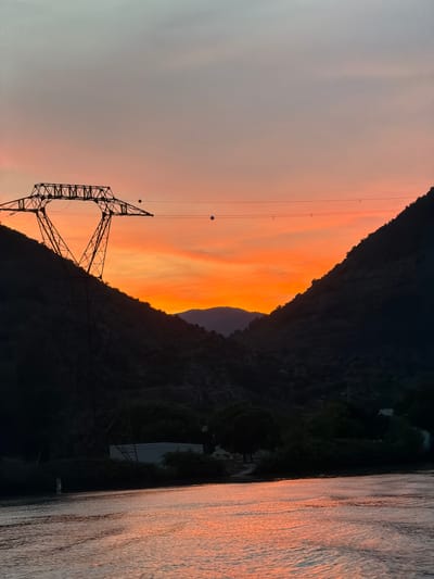 This is a photograph taken in Loriol-sur-Drôme, France. The image depicts a sunset over a body of water, likely a river given the context of the location. The sky is filled with warm hues of orange and pink, indicative of the time of day, which is sunset. In the foreground, a large, dark silhouette of a power line pylon stands prominently against the vibrant sky, with power lines stretching across the frame. These lines are strung between poles that are mostly obscured by the darkness of the mountainside.

The scene is dominated by the stark contrast between the bright, illuminated sky and the dark, mountainous landscape. The mountains themselves are covered in vegetation, creating a textured, dark silhouette. To the right of the frame, the water reflects the colors of the sunset, creating shimmering patterns on its surface. The water appears calm, with gentle ripples disturbing the reflection.

There are no people visible in the image. The overall mood is serene and peaceful, capturing the beauty of a natural sunset amplified by the artificial structures of the power lines. The image does not contain any visible text.
