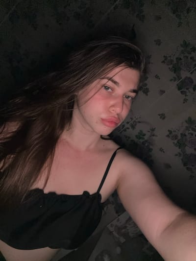 The image shows a young woman lying down. She has long, dark brown hair that is partially covering her face and shoulder. She is wearing a black top with a thin strap. Her eyes are open and she is looking towards the camera, with her lips slightly pursed. The background is dark and appears to have a floral pattern. The lighting is dim, suggesting it might be nighttime or an indoor setting with low light. The overall mood of the image is intimate and relaxed, with the woman seemingly posing for a selfie. There is no visible text in the image. The location context provided is Мещёры, Russia, but there are no specific visual cues in the image to confirm this location.