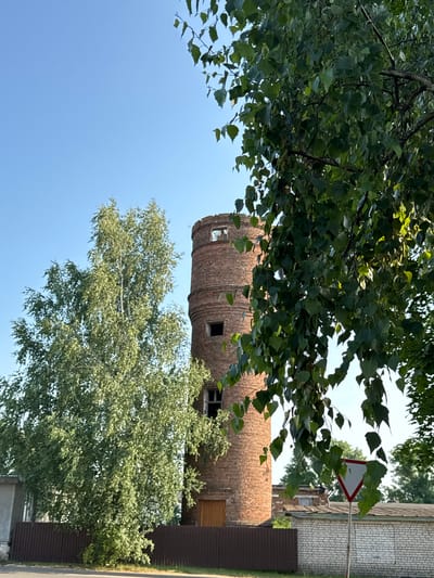 The image shows a cylindrical brick structure with openings, likely an old industrial building or tower, situated in Cherven, Belarus. In the foreground, leafy green trees, possibly birch, with branches extending into the frame, partially obscure the view of the structure. The sky is clear and blue, suggesting a bright, sunny day. To the right of the brick building, a fence and a brick wall are visible, indicating a residential or industrial area. A triangular road sign with a white background and a red border, common in Belarus, is also present, mounted on a pole. The scene appears to be captured in daylight, likely late morning or afternoon, given the quality of light. No people are visible in the frame.