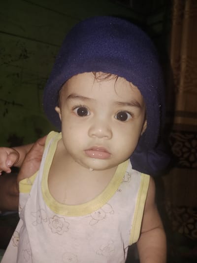 A close-up shot features a young child with dark hair and large brown eyes, wearing a dark blue knit cap and a white and yellow tank top. The child's mouth is slightly open, with a drip of liquid visible on their chin. A hand is visible on the left side of the frame, partially obscuring the child's shoulder. The background consists of a dark wall with faint patterns and a patterned curtain on the right. The scene appears to be indoors. This image was taken in Meunasah Saha, Aceh, Indonesia.