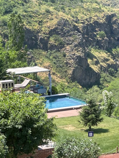 In Byurakan, Armenia, a residential scene features a meticulously maintained outdoor area adjacent to a mountainous landscape. The focal point is a clear blue swimming pool with a wooden deck, surrounded by lush green grass and mature trees. A modern outdoor seating area with a canopy is positioned on the deck, offering a shaded space for relaxation. In the background, steep, rocky hills are covered in dense vegetation, indicating a natural, untouched environment. The scene is bathed in bright sunlight, casting shadows that highlight the contours of the terrain and vegetation. A small, blue sign with a white pictogram of a walking person is visible on the lawn, suggesting the presence of walking paths or regulations.