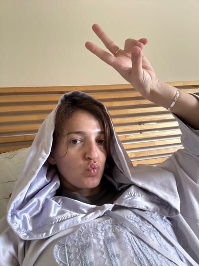 The scene is located indoors, presumably in a bedroom within Anavissos Municipal Unit, Greece. An adult female with short brown hair is centrally positioned, looking directly at the camera. She is wearing a light lavender-colored hooded garment, possibly a bathrobe, featuring a satin-like lining within the hood and intricate lace or embroidered detailing on the chest, secured by visible laces. A dark grey undergarment is partially visible beneath. Her lips are puckered into a "kissing" expression. Her right hand is raised above her head, performing a V-sign gesture with her index and middle fingers extended, while her other fingers are curled. A thin gold-colored ring is present on her right ring finger, and a silver-toned bangle bracelet with embedded patterns is on her right wrist. The background consists of a light beige wall and a light-colored wooden headboard with horizontal slats. A white pillow or bedding is visible on the left side of the frame.