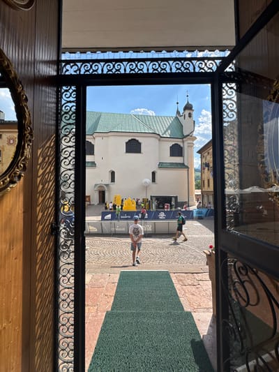 This image captures a view through an open doorway into a town square in Cortina d'Ampezzo, Italy. In the foreground, a green mat leads from the doorway into the square. The square is paved with cobblestones and features a large white building with a green roof, likely a church, in the background. Several people are present in the square, including individuals walking and standing around event banners. The banners suggest some sort of outdoor event or festival. The overall atmosphere is bright and sunny, with a clear blue sky and scattered clouds.