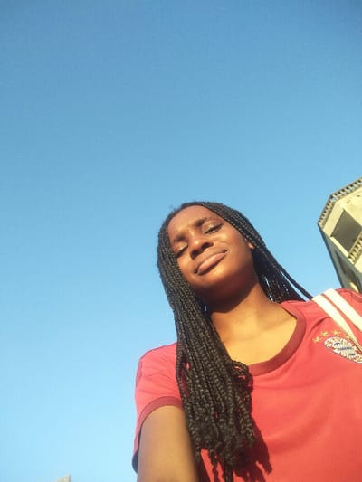 A young adult female with dark skin and long, black box braids is captured in a low-angle outdoor portrait. Her face is tilted upwards, with eyes closed or heavily squinted and lips curved slightly, indicative of a relaxed expression under direct, bright sunlight. She wears a red short-sleeved t-shirt, prominently featuring the FC Bayern Munich club crest on the upper left chest. A light-colored strap, consistent with a shoulder bag, is visible over her right shoulder. The dominant background is a clear, cloudless bright blue sky. In the upper right quadrant of the image, a partial view of a light-colored, angular, multi-story building, constructed of concrete or a similar material, is visible. This structure displays distinct rectangular openings or windows and features architecture consistent with an urban setting in Cotonou, Benin.