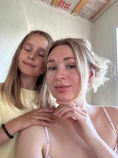 An overhead shot captures two females, one adult and one child, posing for a selfie indoors. The adult, positioned in the foreground, looks directly at the camera, her head tilted slightly, with her right hand touching her chin. The child is behind her, her eyes closed, with a serene expression and her left arm resting on the adult's shoulder. Both individuals have blonde hair. The background shows a wall and a ceiling with exposed wooden beams, suggesting ongoing construction or renovation. The image composition emphasizes the facial features of both subjects and their interaction.