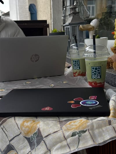 The image depicts an outdoor table scene in Bad Nauheim, Germany, featuring two laptops and two beverages. In the foreground, a closed black laptop with several stickers is positioned horizontally on a light-colored tablecloth patterned with faded yellow roses and green leaves. The stickers include multiple red and orange heart shapes, a circular blue sticker with a cartoon design and partial text "TYKJA", a partially visible "ThinkPad" sticker, and a red "B" sticker. Behind this, an open silver HP laptop, with its lid facing the viewer and the HP logo visible, rests on the same tablecloth.
To the right of the HP laptop, two clear plastic cups with black straws contain layered beverages: a red, possibly strawberry, base topped with a lighter green liquid, likely matcha. Both cups feature a white label with a dark green geometric logo and the text "DOA". The liquid levels indicate partial consumption.
In the mid-ground, a person wearing a light-colored shirt or jacket is partially visible from the back and left shoulder, positioned behind the HP laptop, suggesting engagement with it.
The background shows an outdoor setting. Directly behind the beverages, a gray decorative lantern is visible, followed by a white ceramic duck figurine and potted green plants. Further back, a white building structure, a dark wooden railing or fence, and parts of another building with a window are discernible under an overcast sky.