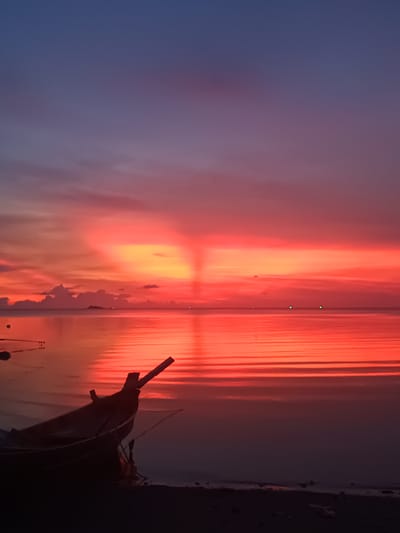 The image depicts a sunset scene in Baan Wok Tum, Thailand, featuring a traditional Thai fishing boat in the foreground. The boat is dark and silhouetted against the vibrant red and orange colors of the sky and its reflection on the water. The sky is a gradient of blue, purple, red, and orange, with a distinct ray of light beaming upward from the horizon. The water is calm and reflects the colors of the sky, creating horizontal bands of red and orange. In the distance, there are a few small islands or landmasses and several dim lights, possibly from boats or structures on the islands. The foreground shows the shoreline, dark and barely visible at the bottom of the image.